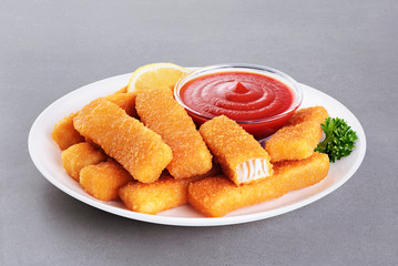 Fish fingers sticks on a plate with parsley, lemon and ketchup on a gray texture.