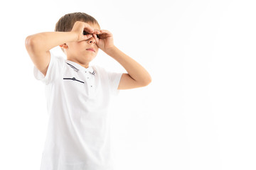 cute boy with bangs in a white t-shirt holds his hands like binoculars on a white background with copy space