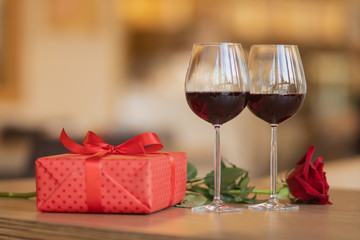 Wine glasses, present and roses on the wooden table in front of beige warm background. Love and holiday concept