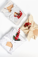 Obraz na płótnie Canvas Christmas gifts wrapped in light textile and decorated Christmas branches with berries, flat lay. Christmas gifts in eco-friendly packaging on a white background, top view