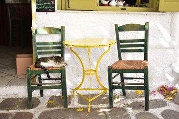 The beautiful holiday island of Ios. A small square in the old town. At a traditional cafe a cat sleeps on a chair in shade away from the summer heat. 