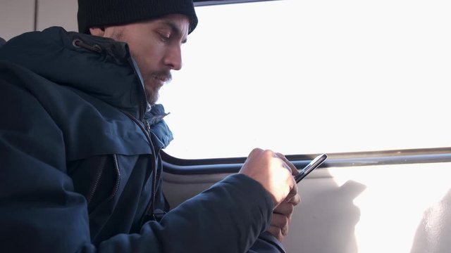 Side view of handsome bearded man in winter parka and knitted hat uses mobile phone while   travelling by train. Millennial male browsing on smartphone in public transport and sneezes.