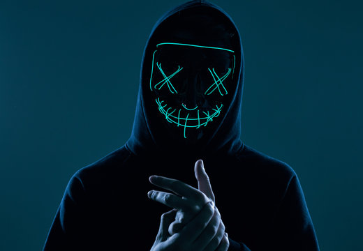 Anonymous man in black hoodie hiding his face behind a neon mask
