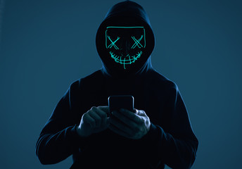 Anonymous man in a black hoodie and neon mask hacking into a smartphone