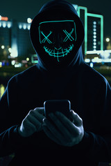 Anonymous man in a black hoodie and neon mask hacking into a smartphone