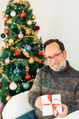 Obraz na płótnie Canvas Portrait of middle age man posing next to Christmas tree, wearing warm pullover and glasses, holding a white box gift with red ribbon