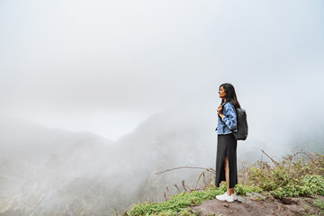 Young woman tourist with backpack relaxing on top of a mountain and enjoying the view of the misty valley