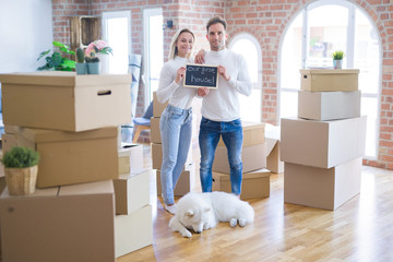 Fototapeta premium Young beautiful couple with dog standing holding blackboard with message at new home around cardboard boxes