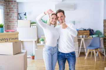 Young beautiful couple standing at new home around cardboard boxes smiling making frame with hands and fingers with happy face. Creativity and photography concept.