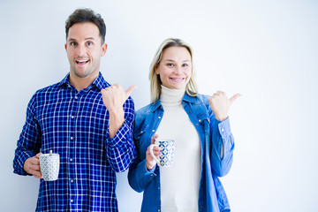 Young beautiful couple drinking cup of coffee standing over isolated white background pointing and showing with thumb up to the side with happy face smiling