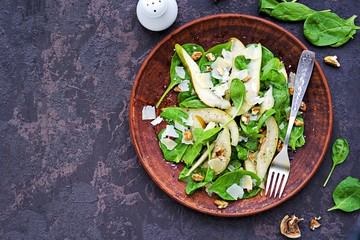 Green salad of fresh spinach, juicy pear, walnuts and parmesan on a brown clay plate on a dark concrete background. Vegan recipes. Healthly food. Copyspace.