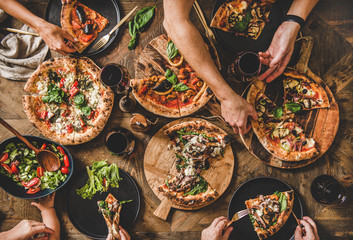 Family or friends having pizza party dinner. Flat-lay of people eating different kinds of Italian pizza, salad and drinking wine over wooden table, top view. Fast food lunch, gathering, celebration