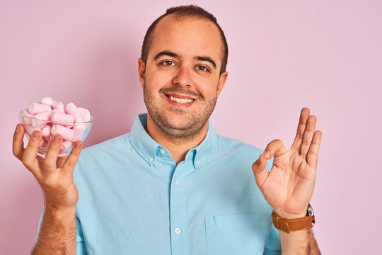 Young man holding bowl with marshmallows standing over isolated pink background doing ok sign with fingers, excellent symbol