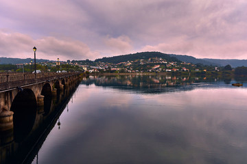 Obraz premium Ancient Roman stone bridge over calm waters and in the background typical village of northern Spain on hill and reflection in the water.