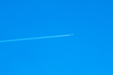 Plane flies high in clear blue sky from left to right, leaving jet trail behind