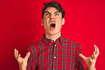 Teenager boy wearing red shirt standing over isolated background crazy and mad shouting and yelling with aggressive expression and arms raised. Frustration concept.