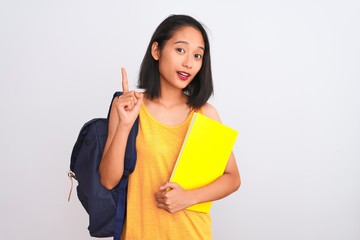 Young chinese student woman wearing backpack holding book over isolated white background surprised...