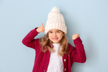 Cute girl in winter clothes showing thumb-up on color background