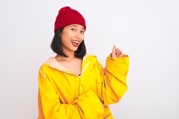 Young beautiful chinese woman wearing raincoat and wool cap over isolated white background with a big smile on face, pointing with hand and finger to the side looking at the camera.