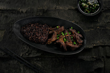  Delicious sticky Asian beef meat in black pepper sauce with black rice on a rustic wooden black table. Directly above top view shot.