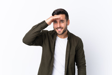 Young handsome man with beard over isolated white background looking far away with hand to look something