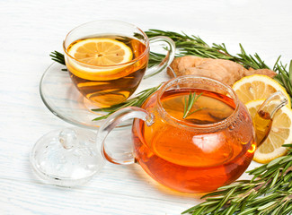 Glass teapot and glass cup of tea with fresh rosemary branches, ginger roots and lemon on a white wooden background. 