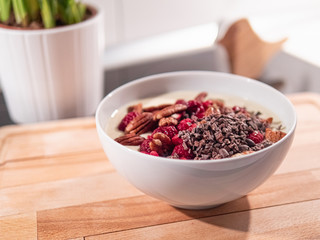 Eine weiße Keramikschüssel mit Joghurt, Pecannüssen, Kakaonibs und Himbeeren, steht in der Küche auf einem hellen Holzbrett.