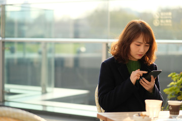 One Asian beautiful young woman sitting outdoor under sunshine, tapping on smartphone. Blur background