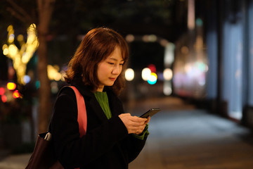 One Asian young woman texting message on phone outdoor at night. Blur colorful street lights background