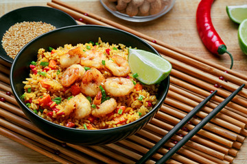 Delicious fried rice with shrimps, lime and vegetables in a black bowl on a bamboo placemat. Traditional Asian food on a wooden table.