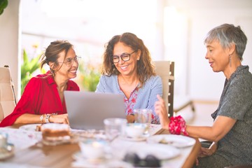 Meeting of middle age women having lunch and drinking coffee. Mature friends smiling happy using laptop at home on a sunny day