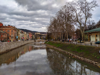 Fototapeta na wymiar Beautiful view of Miljacka river in Sarajevo, Bosnia on a cloudy autumn day. Historical buildings and stone bridges in old part of the town.