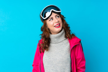 Skier woman with snowboarding glasses over isolated blue wall laughing and looking up