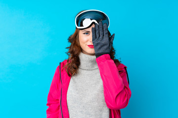 Skier woman with snowboarding glasses over isolated blue wall covering a eye by hand