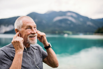 Senior man with earphones standing by lake in nature, listening to music.