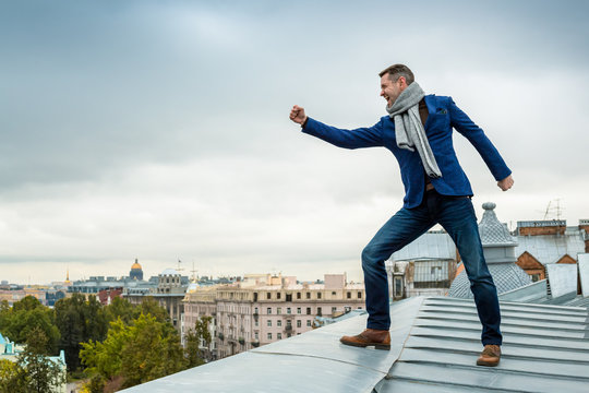 An emotional stylish young man in jeans, a blue jacket and a scarf stands on the edge of the roof of the building. The person shows a hand gesture with a clenched fist. Saint-Petersburg, Russia.