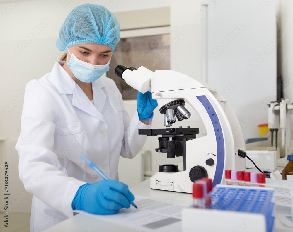 Poster cheerful girl assistant working with microscope and writing results of research