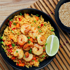 Quick and healthy Asian meal made of fried rice, fresh shrimps, lime and vegetables in a black bowl on a wooden table. Top view, directly above shot with space for text.