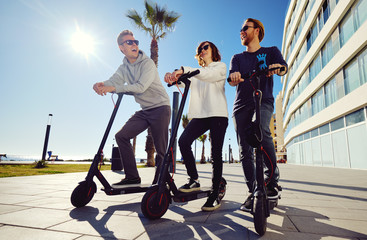 Obraz premium Three best friends young 20s -30s girl and guys spend time outdoors gathered together driving on electric scooter modern land vehicle, easy comfort usage, technology urban transportation concept image
