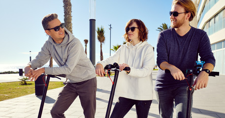 Three best friends young 20s -30s girl and guys spend time outdoors gathered together driving on electric scooter modern land vehicle, easy comfort usage, technology urban transportation concept image