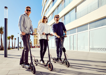 Three best friends young 20s -30s girl and guys spend time outdoors gathered together driving on electric scooter modern land vehicle, easy comfort usage, technology urban transportation concept image
