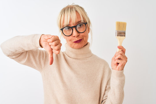 Middle age painter woman holding paint brush standing over isolated white background with angry face, negative sign showing dislike with thumbs down, rejection concept