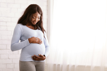 Peaceful pregnant afro woman hugging her tummy at home
