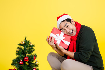 Happy Asian man wearing green sweater and Santa hat holding gift box sitting on yellow background.
