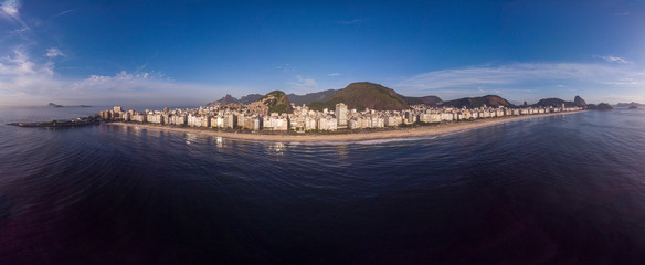 Full aerial view of Copacabana beach and neighbourhood at early sunrise flanked by the Sugarloaf mountain on the right and Copacabana fort on the left against a blue sky seen from the ocean