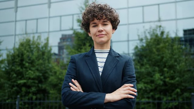 Portrait of attractive ambitious businesslady standing outside with arms crossed with light smile on her face looking at camera, business center is in background.