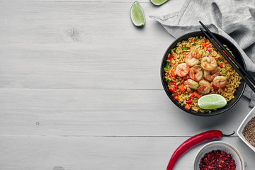 Delicious fried rice with shrimps, lime and vegetables in a black bowl. Traditional Asian food on a white wooden table with a fabric tablecloth. Top view, directly above shot with space for text.