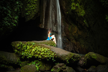 Young Caucasian woman sitting on the rock and enjoiyng waterfall landscape. Travel concept. Leke Leke waterfall, Bali.