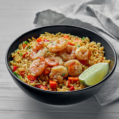 Delicious fried rice with shrimps, lime and vegetables in a black bowl. Traditional Asian food on a white wooden table with a fabric tablecloth.