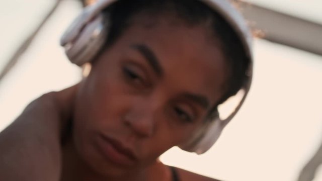 Close up view of serious sporty african american woman with wireless headphones touching her neck while sitting on the old bridge